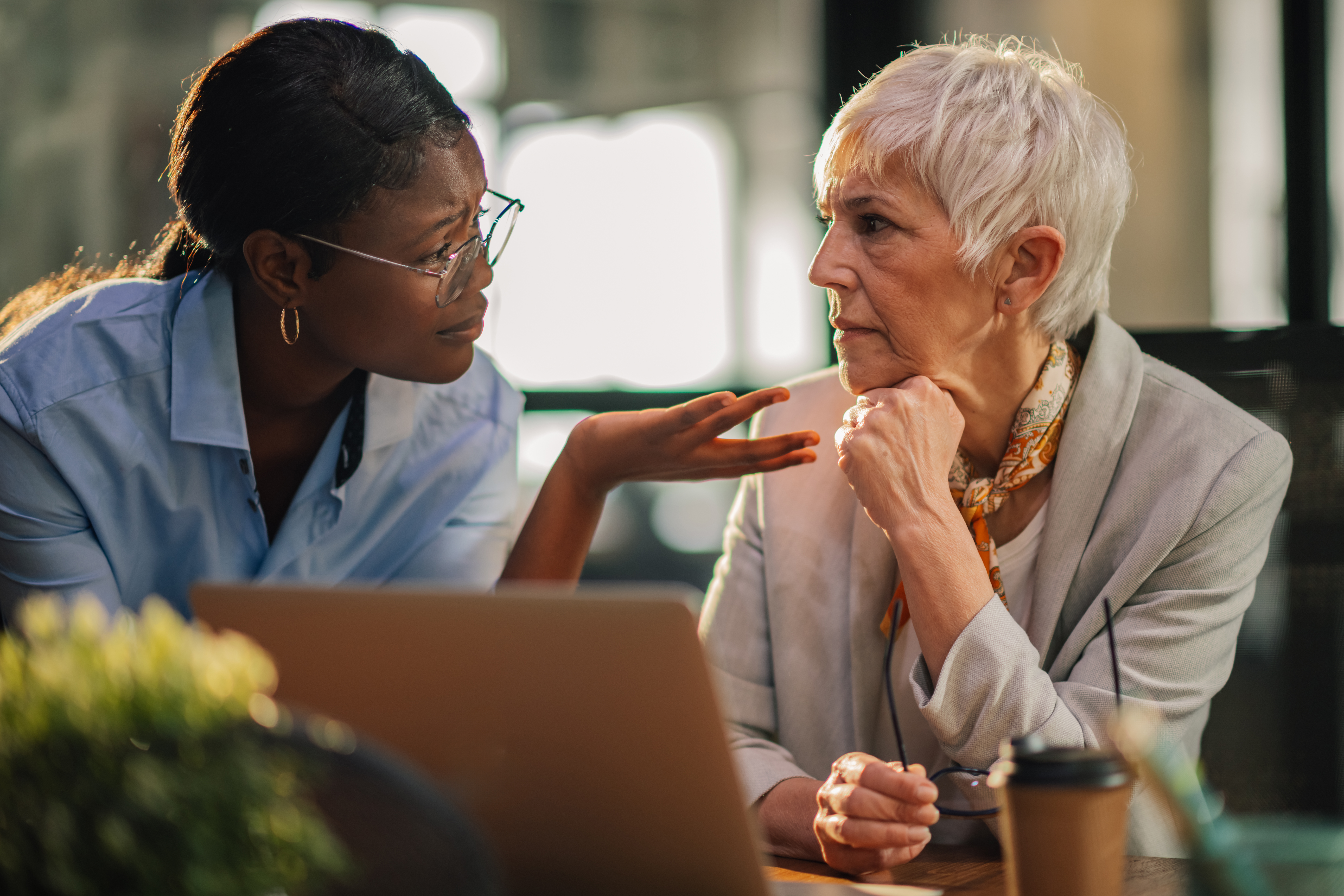 African american office worker explaining and discussing business strategy with her mature female mentor at modern workspace. Multicultural businesswomen having consultations about startup project.